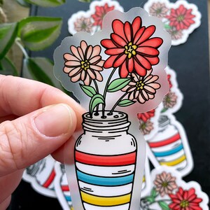 May include: A clear glass jar with red, blue, and yellow stripes, filled with a bouquet of pink and red flowers. The jar has a white lid and is decorated with a simple floral design.