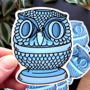Pode incluir: Um vaso de cerâmica azul em forma de coruja com uma superfície texturizada. O vaso tem uma base larga e uma parte superior redonda. Os olhos da coruja são grandes e redondos, e o bico é pequeno e pontudo.