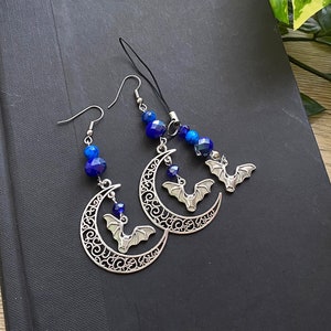 Op de afbeelding: Een paar zilveren oorbellen met een halve maan ontwerp en een vleermuis bedel. De oorbellen zijn versierd met blauwe glazen kralen.