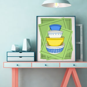 Może przedstawiać: Wydruk w ramie przedstawiający stos czterech kolorowych misek z niebieskimi i białymi wzorami na zielonym tle. Miski są ułożone w kształcie piramidy, z największą miską na dole i najmniejszą na górze. Wydruk wisi na ścianie nad jasnoniebieskim biurkiem z dwoma szufladami i różową podstawą.