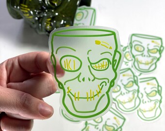 Op de afbeelding: Groene en gele sticker met een cartoon schedel gezicht. De schedel heeft genaaide ogen en mond. De sticker is op een witte achtergrond.