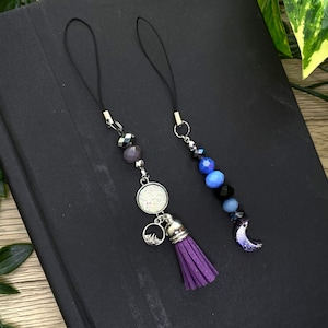 May include: Two phone charms with a black cord and silver accents. One charm features a purple tassel and a silver charm with a mountain design. The other charm features a crescent moon charm with blue and black beads.