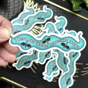 May include: A collection of stickers with a teal banner design. The banner reads "I kept the inside thoughts inside" in white text, with bat and sword illustrations. The stickers are on a black surface.