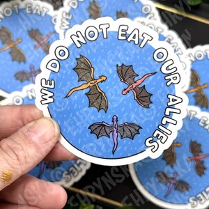 May include: A round sticker with a blue background and the text "WE DO NOT EAT OUR ALLIES" in white. The sticker features three cartoon dragons in orange, pink, and purple.
