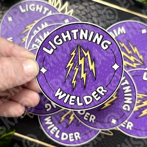May include: A hand holding a purple sticker with the words "LIGHTNING WELDER" in white, and a yellow lightning bolt graphic. Additional stickers are visible in the background.