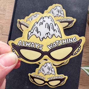 May include: A stack of yellow stickers with a cat-eye sunglasses design and the text "ALWAYS WATCHING". The sunglasses have black frames and a pearl-like detail. The top of the sticker features a mountain range.