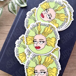 May include: A stack of round stickers featuring a cartoon character with yellow hair and red lips. The stickers have a light green background and the text "JUST A HOT MESS EXPRESS" in a circular pattern.