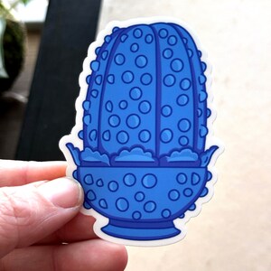 May include: A blue and white sticker of a cartoon cactus with a pot. The cactus is covered in small blue circles and has a white outline. The pot is also blue and has a white outline.