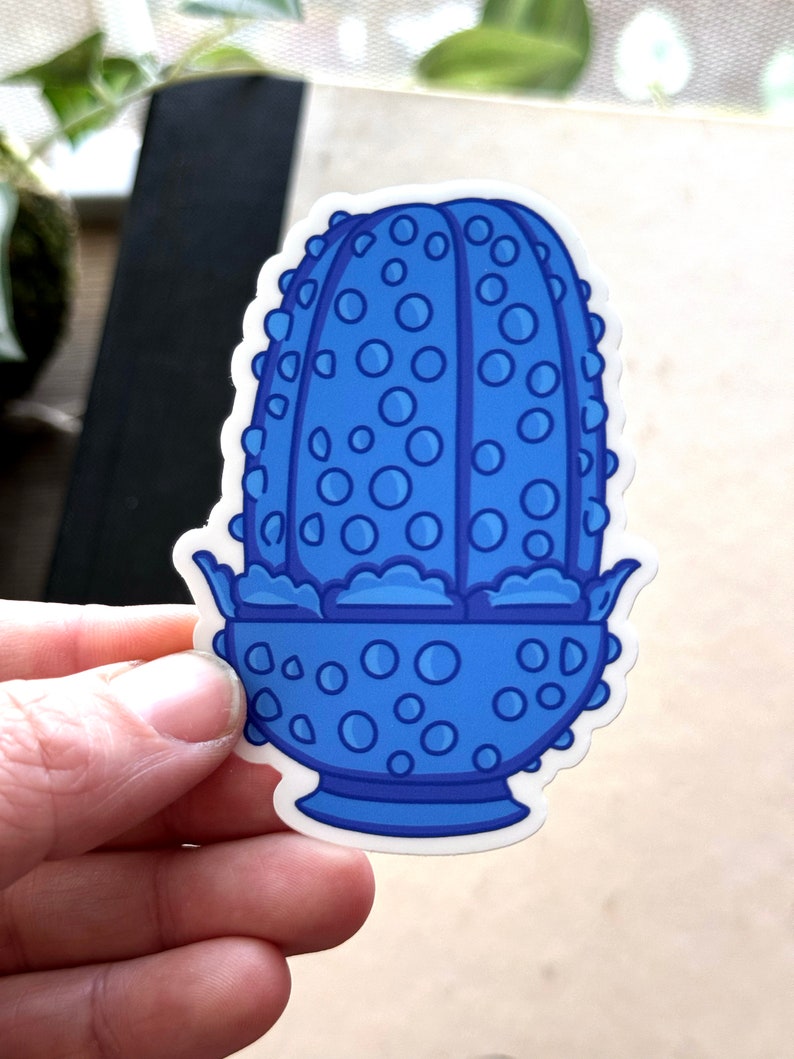 May include: A blue and white sticker of a cartoon cactus with a pot. The cactus is covered in small blue circles and has a white outline. The pot is also blue and has a white outline.