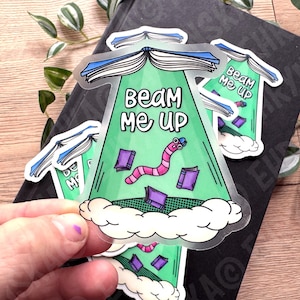 May include: A set of transparent stickers with a whimsical design. Each sticker features a green beam with the text "BEAM ME UP", a cartoon worm, and floating books. The stickers are on a black surface.