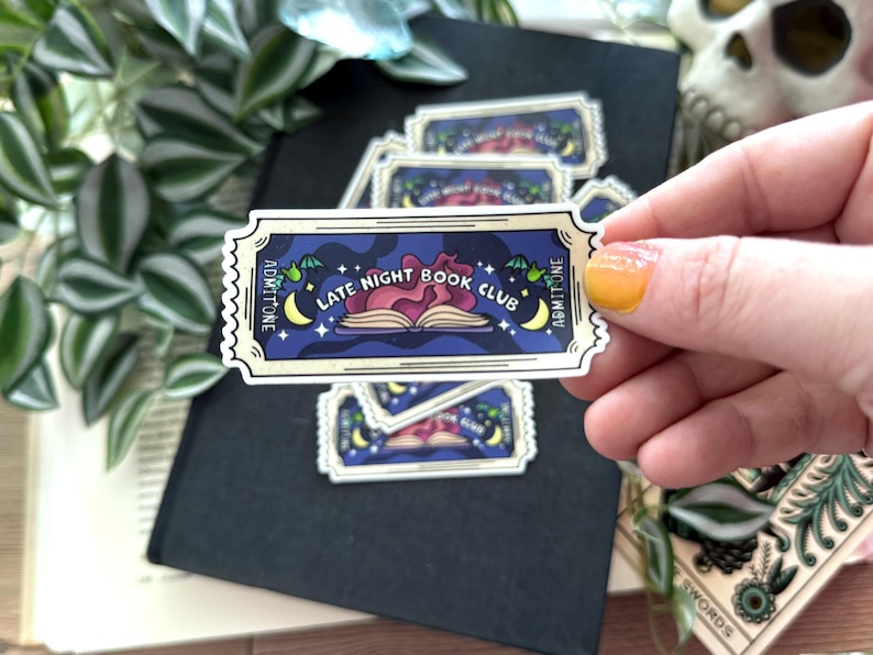 Peut inclure: Un autocollant avec un design de billet vintage repr&eacute;sentant un livre avec un c&oelig;ur rouge incandescent et le texte "Late Night Book Club". L'autocollant a un fond bleu avec des &eacute;toiles et des croissants de lune.