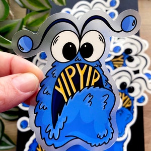 Könnte beinhalten: Ein blaues, flauschiges Monster mit großen Augen und einem weit geöffneten Mund. Der Mund des Monsters ist offen und das Wort "YIP" ist darin sichtbar. Das Monster ist ein Aufkleber.