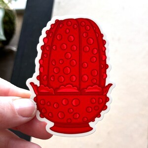 May include: A red, cartoon-style, ceramic planter with a textured, dotted surface. The planter has a wide, round base and a narrow, cylindrical top.