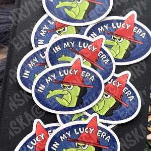 Peut inclure: Plusieurs autocollants ovales avec une sorci&egrave;re de dessin anim&eacute; &agrave; la peau verte, un chapeau rouge et le texte "IN MY LUCY ERA". La sorci&egrave;re a un long nez et une expression s&eacute;v&egrave;re. Le fond est bleu fonc&eacute; avec des &eacute;toiles blanches.