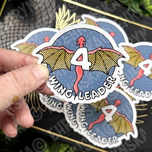 May include: A sticker featuring a red dragon with yellow wings and the number 4. The text "WING LEADER" is written around the dragon in a circle. The sticker is held by a hand.