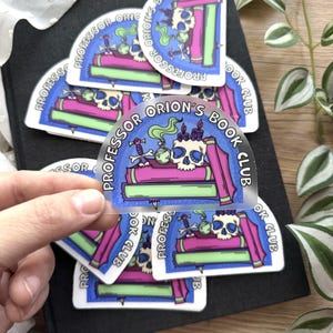 May include: A collection of semi-transparent stickers with a blue border and the text "PROFESSOR ORION'S BOOK CLUB". Each sticker features a skull, books, and potion bottles in pink, green, and purple. The stickers are on a black book.