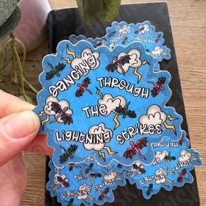 May include: A die-cut sticker with a light blue background featuring white clouds, yellow lightning bolts, and small flying dragons in various colors. The text reads Dancing Through The Lightning Strikes in a playful, hand-lettered black font.