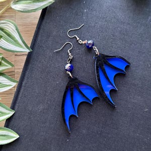 Op de afbeelding: Een paar blauwe en zwarte acryl oorbellen in de vorm van vleugels. De oorbellen hebben elk een donkerblauwe kristallen kraal.