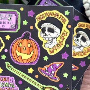 Peut inclure: Planches d'autocollants noirs avec des illustrations sur le th&egrave;me d'Halloween. Les motifs comprennent une citrouille souriante, un cr&acirc;ne portant un chapeau avec le texte "SEE YOU IN THE AFTERLIFE!", un chapeau de sorci&egrave;re et un balai. Les autocollants sont entour&eacute;s d'&eacute;toiles et de points.