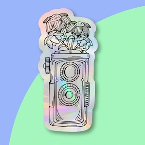 May include: A holographic sticker featuring a vintage camera with a bouquet of flowers inside. The camera has two lenses and a winding knob. The flowers are stylized and have a simple design.
