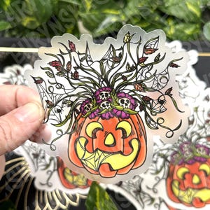May include: A clear sticker featuring a smiling jack-o'-lantern with a spider web design. The pumpkin is orange and yellow, with a floral arrangement of skulls and leaves. The sticker is held by a hand.