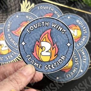 May include: A collection of round stickers featuring a flame design with the text "Fourth Wing Flame Section 2". The stickers have a blue background with white text and a stylized flame in red, orange, and yellow. The stickers are held by a hand.