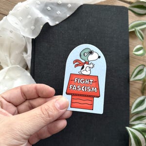 May include: A sticker featuring Snoopy in a pilot's helmet and scarf, standing on his doghouse. The doghouse has the words "FIGHT FASCISM" in red letters. The sticker is held in a hand.