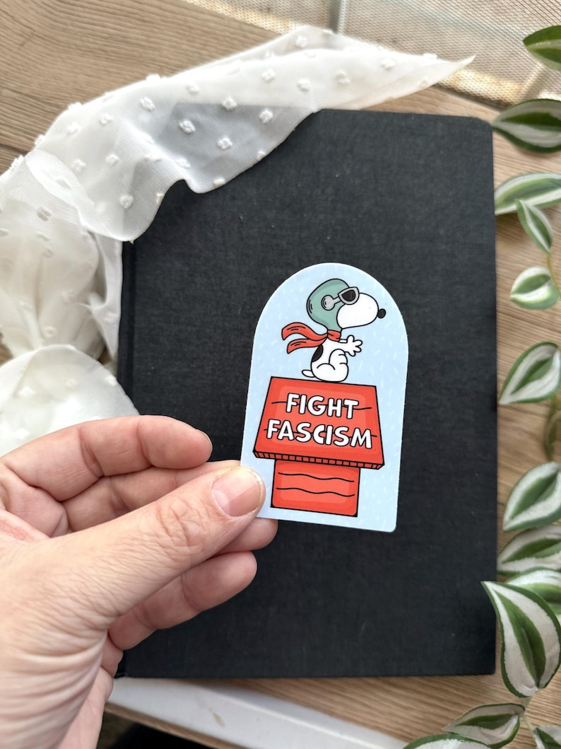 May include: A sticker featuring Snoopy in a pilot's helmet and scarf, standing on his doghouse. The doghouse has the words "FIGHT FASCISM" in red letters. The sticker is held in a hand.