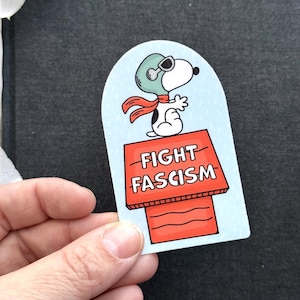 May include: A sticker featuring Snoopy in a pilot's helmet and scarf, standing on a red doghouse. The doghouse has the words "FIGHT FASCISM" in white. The background is light blue.