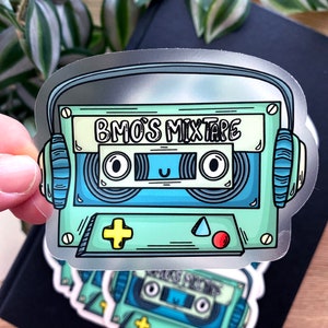 Puede incluir: Una pegatina plateada de una cinta de casete de dibujos animados con auriculares. La cinta de casete tiene un diseño verde y azul con el texto "BMO'S MIXTAPE" en la etiqueta.