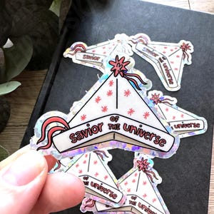 Puede incluir: Pegatinas holográficas con un diseño triangular blanco, detalles en rojo y el texto "Savior of the Universe". Las pegatinas tienen un borde rosa y un diseño de estrella.