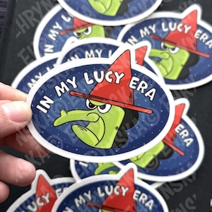 Peut inclure: Plusieurs autocollants ovales avec une sorci&egrave;re de dessin anim&eacute; verte, un chapeau rouge et le texte "IN MY LUCY ERA" en blanc. La sorci&egrave;re est sur un fond bleu fonc&eacute; &eacute;toil&eacute;. Plusieurs autocollants sont visibles.