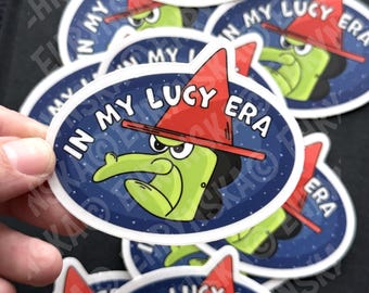 May include: A collection of oval stickers. Each sticker features a cartoon witch with green skin, a red hat, and the text "IN MY LUCY ERA" in white. The witch is set against a dark blue starry background. Multiple stickers are visible.