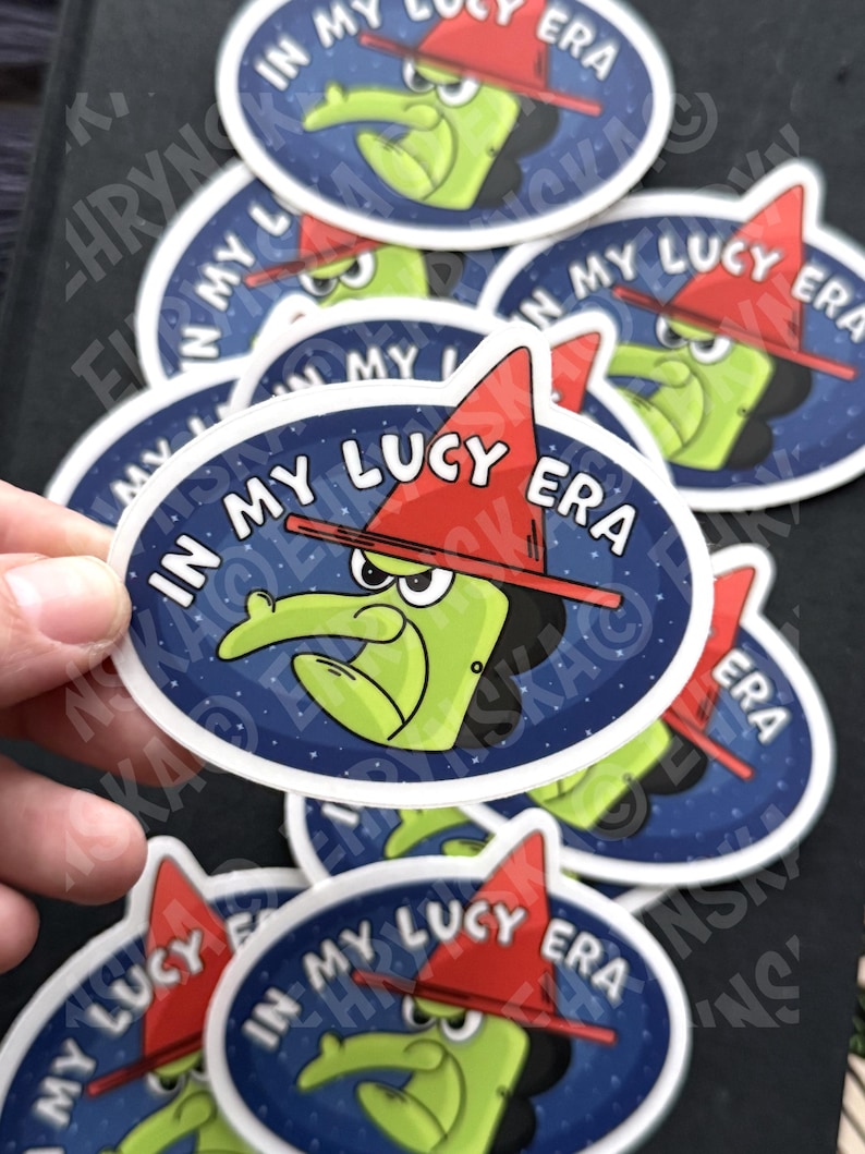 Peut inclure: Plusieurs autocollants ovales avec une sorci&egrave;re de dessin anim&eacute; verte, un chapeau rouge et le texte "IN MY LUCY ERA" en blanc. La sorci&egrave;re est sur un fond bleu fonc&eacute; &eacute;toil&eacute;. Plusieurs autocollants sont visibles.