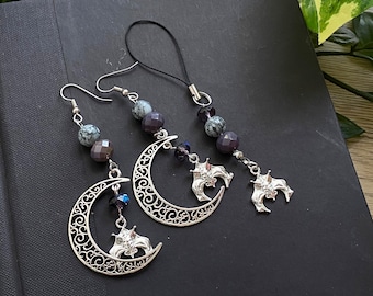 Puede incluir: Un conjunto de tres pendientes de plata con charms y un charm para teléfono con forma de luna creciente con un murciélago colgando. Los charms están decorados con cuentas de color gris oscuro y blanco moteado y cristales azul oscuro.