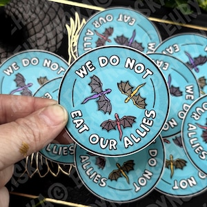 May include: A collection of round stickers with a light blue background and the text "WE DO NOT EAT OUR ALLIES". Each sticker features three cartoon dragons in purple, yellow, and pink.