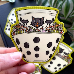 Könnte beinhalten: Eine weiße Keramikschüssel mit schwarzen Punkten, gefüllt mit Candy Corn und anderen Halloween-Süßigkeiten. Eine schwarze Katze mit grünen Augen schaut aus der Schüssel.
