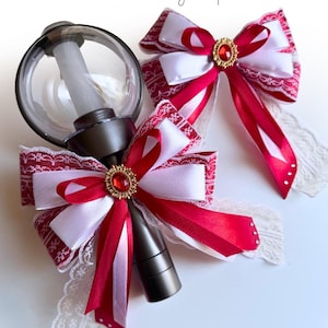May include: A dark gray light stick with a clear globe top, adorned with a decorative bow featuring red and white satin ribbons, white lace trim, and a central gold-toned oval brooch with a red gem. A matching bow is positioned to the right.