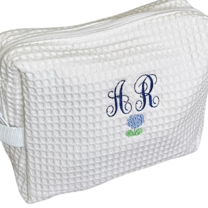 Puede incluir: Neceser blanco con textura de gofre, cierre de cremallera y asas laterales pequeñas. La bolsa presenta las iniciales bordadas "A R" en azul marino, junto con un diseño floral azul. Diseñado para guardar artículos de aseo y objetos personales.