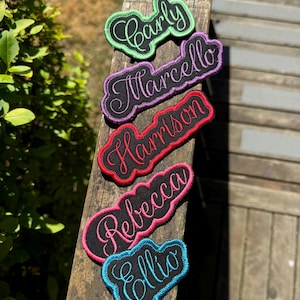 Peut inclure: Cinq écussons brodés avec des noms de différentes couleurs. Les noms Carly, Marcello, Harrison, Rebecca et Ellio sont affichés en écriture cursive sur un tissu noir avec des bordures colorées. Les écussons sont disposés sur une surface en bois.