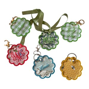 May include: A collection of five scalloped fabric keychains featuring various patterns and embroidery. Designs include green gingham, pink strawberry print, blue bird print, yellow bee print, and a green ribbon-tied tag with the text MOM.