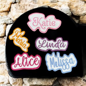 May include: A black object displays embroidered name patches. The names "Katie" in pink, "Maria" in orange, "Linda" in purple, "Alice" in pink, and "Melissa" in blue are stitched onto white cloud-shaped patches with colored borders. The names are in a cursive font.