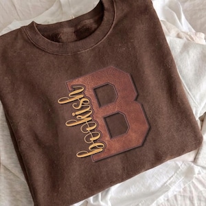 Könnte beinhalten: Ein dunkelbraunes Sweatshirt mit Rundhalsausschnitt und einem großen, strukturierten braunen Buchstaben B in Lederoptik. Das Wort bookish ist in einer kursiven, goldgelben Schrift über die linke Seite des Buchstabens B auf der Vorderseite gedruckt.
