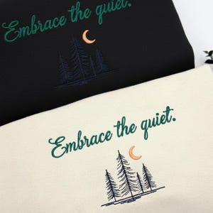 May include: Black and cream-colored sweatshirts with embroidered forest and moon designs. The text "Embrace the quiet" is written in green script above the design. The sweatshirts are folded and stacked.