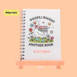May include: A spiral-bound book journal with a white cover featuring a cartoon duck carrying books. The text reads "OOOPS, I BOUGHT ANOTHER BOOK" and "BOOK JOURNAL". The journal is displayed on a small wooden easel.