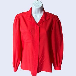 May include: A bright red long-sleeve blouse with a collared neckline and a single pocket on the left side. The blouse is displayed on a white mannequin and features a button-down front. The sleeves have a slight puff at the shoulders.
