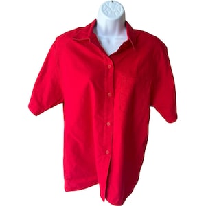 May include: A bright red short-sleeved button-down shirt with a collar and a single chest pocket. The shirt is displayed on a white mannequin. The fabric appears to be a lightweight cotton or blend.