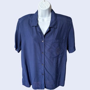 May include: A navy blue short-sleeved button-down shirt with a chest pocket and embroidered emblem. The shirt is displayed on a white mannequin. The fabric appears to be lightweight and has a slight sheen.