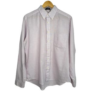 May include: A long-sleeved button-down shirt with vertical stripes in shades of pink and white. The shirt has a button-down collar, a chest pocket, and is hanging on a wooden hanger. The shirt is a classic style.