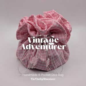 May include: A pink and white gingham dice bag with a drawstring closure. The handmade bag has eight pockets. Text on the bag reads "For the Vintage Adventurer" and "Handmade 8-Pocket Dice Bag". The Thrifty Adventurer is also printed on the bag.